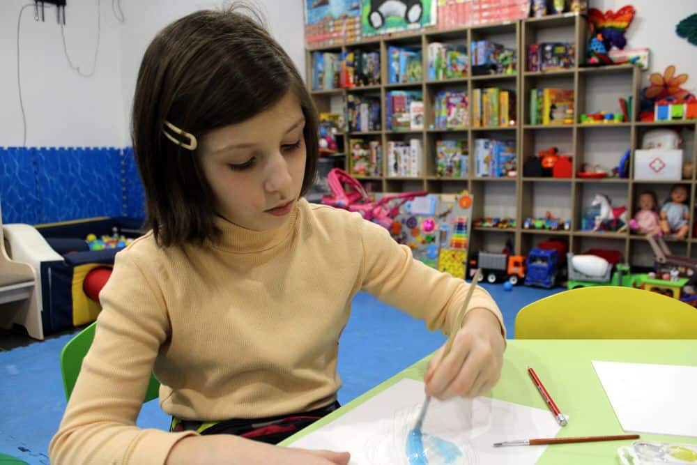 A girl sits at a yellow table and paints on a white piece of paper.