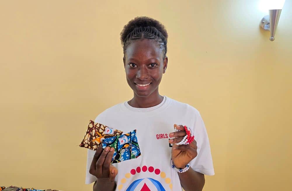 A girl smiles and holds up sanitary pads and beaded bracelets.