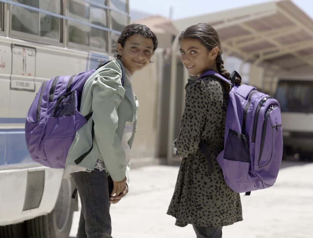 Two girls stand next to a bus wearing purple backpacks.