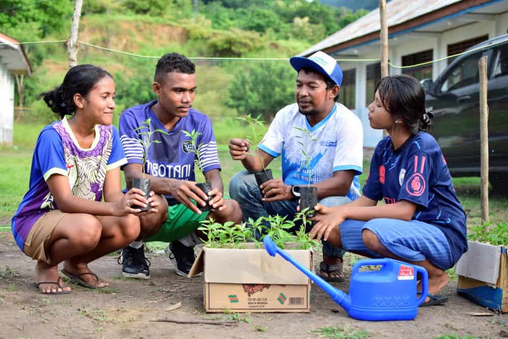 Four young people crouch down in front of a box of plants and watering can.