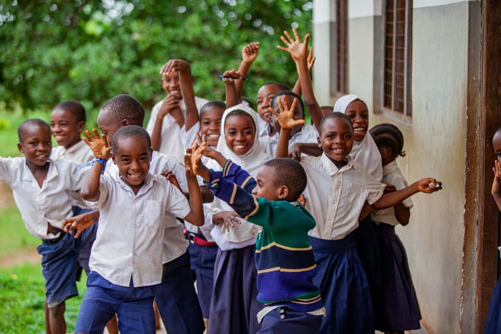 A group of boys and girls smile and wave outside of a school building.