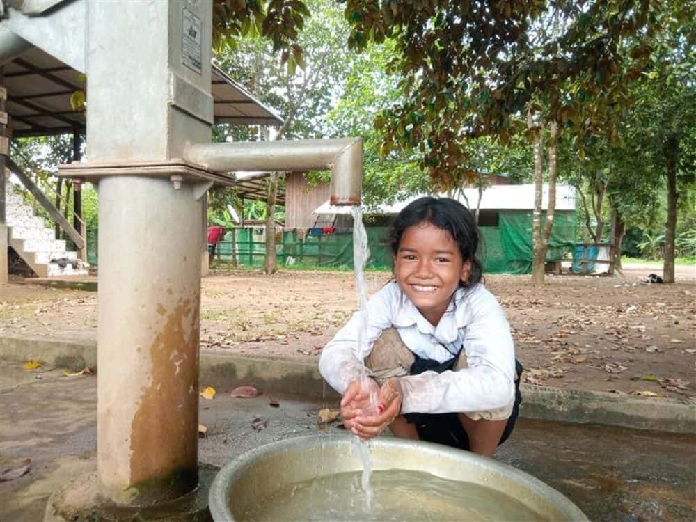 A girl holds her hands out under a water tap and smiles.