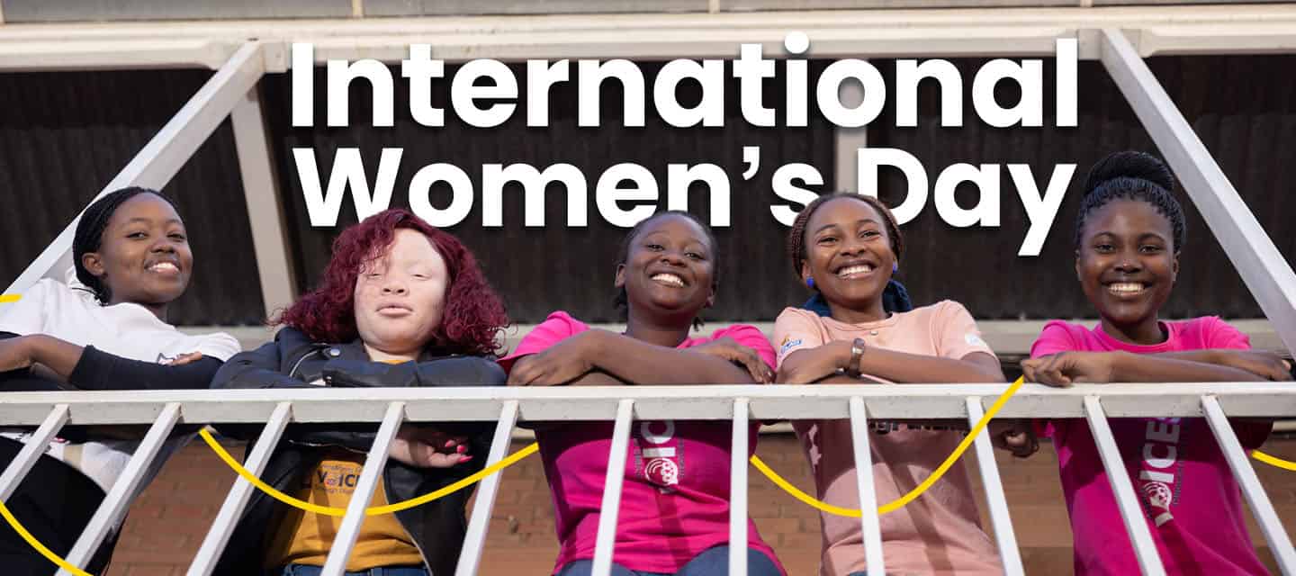 Five teenage girls lean over a balcony railing and smile, with “International Women’s Day” text across the top.