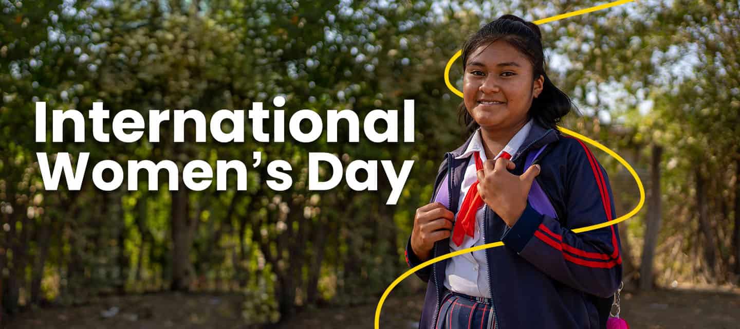 A teenage girl in a school uniform stands outdoors, smiling, with International Women’s Day text on the left.