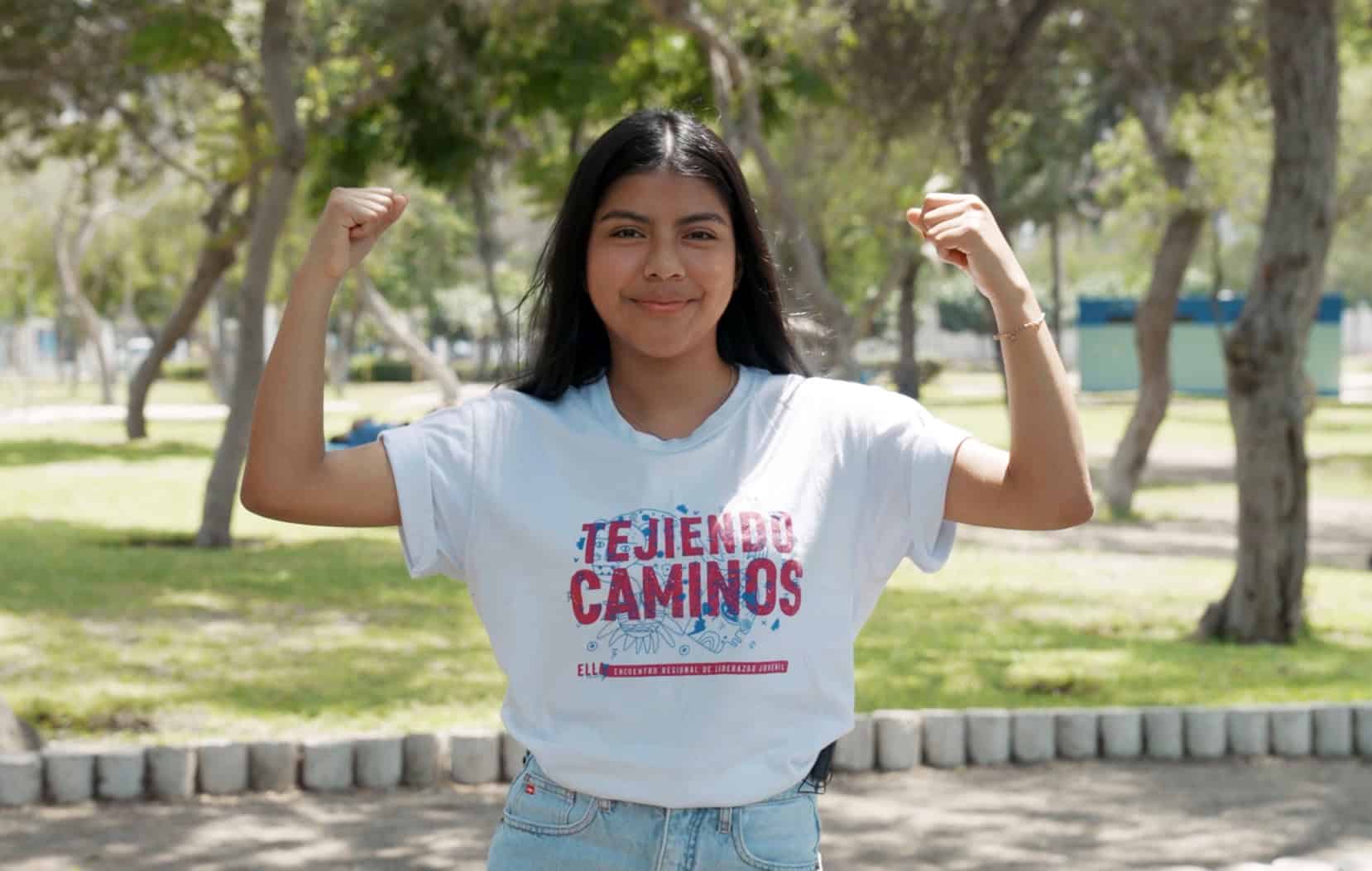 Fernanda, 17, raises her fists in the air outdoors in Lima, Peru, wearing a white Plan International ELLA project T-shirt.
