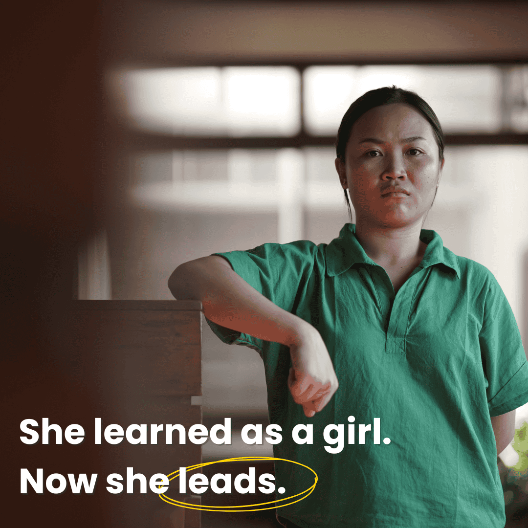 A woman in a green shirt stands indoors, resting her arm on a wooden structure, looking directly at the camera. White text overlay reads, “She learned as a girl. Now she leads.”