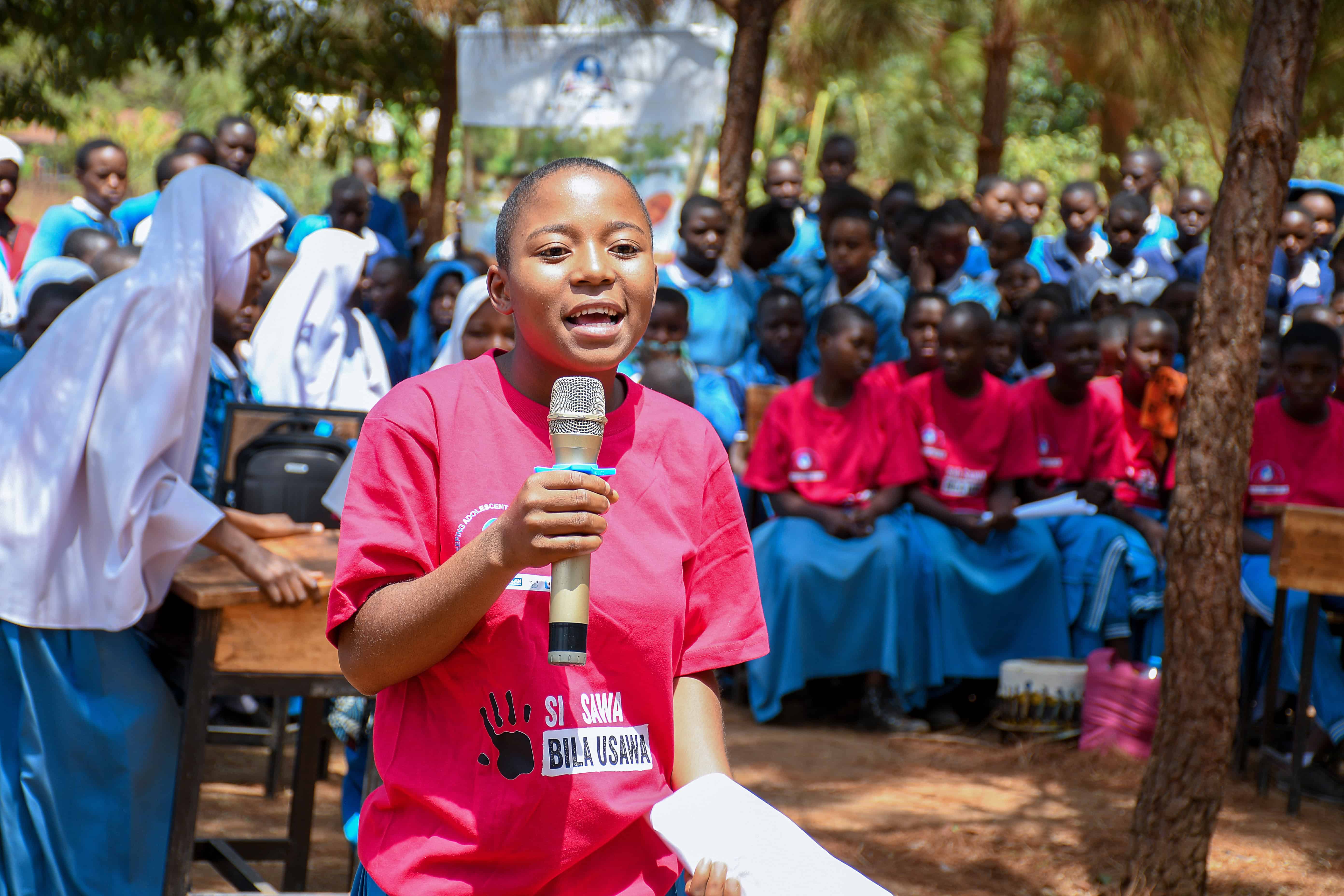 A girl addresses her peers during a community event in Tanzania, wearing a KAGIS T-shirt reading “Si Sawa Bila Usawa.”